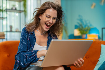 Surprised happy young woman in eyeglasses using laptop computer, receive good news message, mouth...