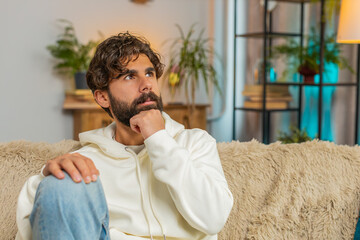 Sad lonely man sits at home room looks pensive thinks over life concerns or unrequited love suffers...