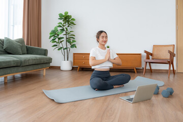 Young Asian woman meditating in lotus position at home, sitting on the floor in fitness clothing. Breathing workout to achieve relaxation, peace and mindfulness