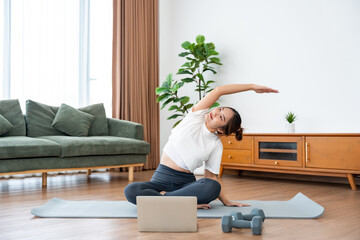 Young Asian woman stretching yoga workout on exercise mat while online training class with computer laptop in living room