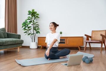Young Asian woman stretching yoga workout on exercise mat while online training class with computer laptop in living room