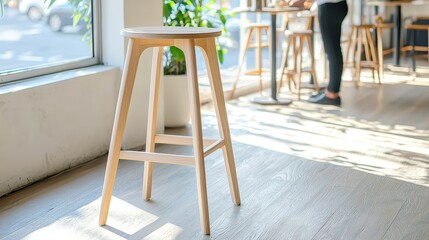 A stylish wishbone stool stands in a sunlit cafe near a barista serving coffee, showcasing its elegant design and light wood finish perfect for modern interiors.