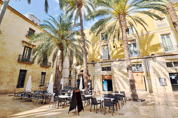 Charming outdoor cafe, nestled in cozy square surrounded by traditional Mediterranean architecture, shaded by tall palm trees. Concept of Mediterranean lifestyle
