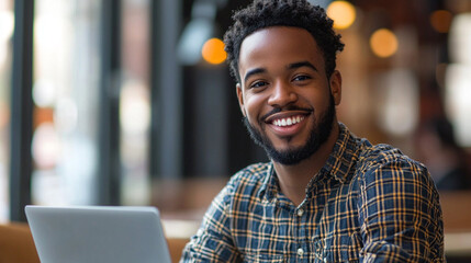 A joyful man is sitting at a cafe, smiling broadly while using his laptop. The warm atmosphere is enhanced by soft lighting and wooden decor, creating a welcoming environment for work