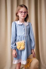 A young girl with long blonde hair stands on fabric background wearing a velvet blue dress with a white collar. A seven year old child wears glasses with pink frames. She gazes thoughtfully forward