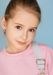 Close-up portrait of a cute girl with light brown hair combed back, wearing a pink shirt and light overalls. She has a gentle expression. The background is soft blue, creating a calm atmosphere.