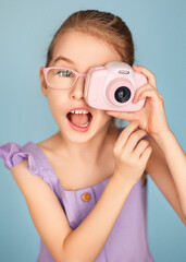 A little girl in a purple top acts like a professional photographer and pretends to be a paparazzi, taking photos with a toy pink camera on a blue background. She opened her mouth in surprise.        