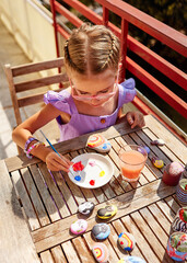 A girl with braided hair paints with acrylic paints on sea pebbles. On the table in front of her are stones that she has already painted during her vacation. She sits and decorates natural materials