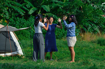 The three girls raise their hands in joy, smiling and celebrating their teamwork after finishing the tent setup in a natural setting.