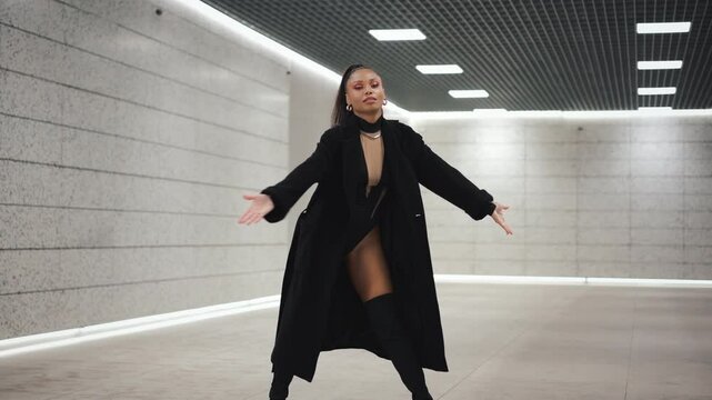 Confident sexy female dancer in black bodysuit and thigh high boots performs stylish choreography in futuristic underground tunnel with minimalistic concrete and LED lighting design