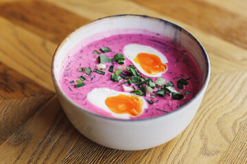 Traditional cold beet soup. Chilled summer dish with eggs. Pink creamy soup in bowl. Rustic wooden table. Fresh herbs topping. Healthy lunch idea. Polish cuisine concept. Chlodnik soup closeup.