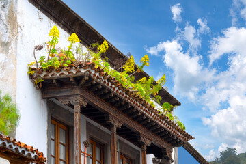 An ornately carved wooden balcony of an old house with lush greenery and yellow flowers growing on the roof.
