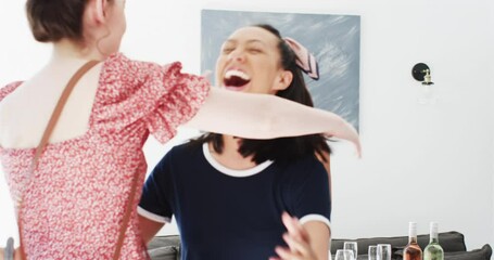 Diverse female friends placing wine glasses at home, hugging arriving friend, reaching bottles - Powered by Adobe