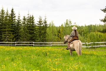 Cowgirl riding on horse (Icelandic horse). Green meadow and trees