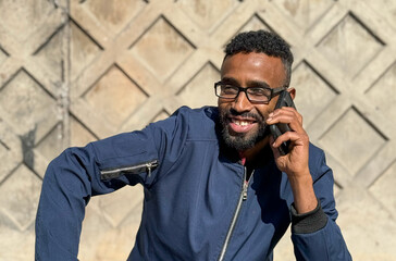 African American man in a blue jacket stands outside of an brick wall, talking on his phone. He...