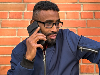 African American man in a blue jacket stands outside of an brick wall, talking on his phone. He...