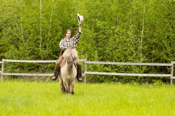 Cowgirl riding on horse (Icelandic horse). Green meadow and trees