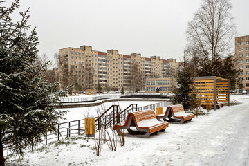 A snow-covered city park with benches and a fir tree against the backdrop of a typical Soviet residential building on a cloudy winter day.