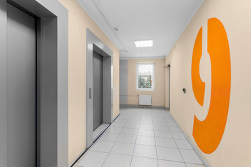 A spacious hall with elevators, a window at the end of the corridor, and a large orange number nine on a light wall.