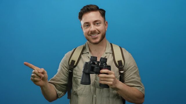 Young man with beard and backpack holding binoculars smiles and points against a blue background, suggesting exploration and travel.