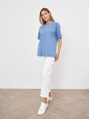 Woman stands confidently wearing a blue shirt and white pants with matching sneakers. The room has a herringbone-patterned wooden floor. Her relaxed posture exudes casual elegance