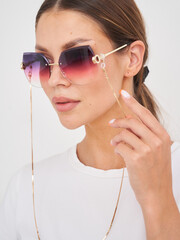 Woman wearing angular gradient sunglasses with a delicate gold chain, holding it lightly. Her hair...