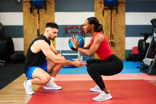 Determined woman performing squats with medicine ball under supervision of personal trainer in modern gym