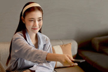 Obraz premium Happy middle aged female holds remote control to switch channels while watches TV show while sits on sofa at living room. Beautiful adult Asian woman spending time while watching movie at home.