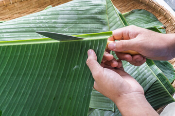 Hands cutting banana leaf with knife for traditional food wrapping, a common eco-friendly practice in Malaysian and Southeast Asian cuisine preparation.