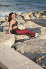 A slightly overweight young woman dressed in sportswear, sitting on a large rock looking at the camera with the evening light shining on her face and the waves breaking on the shore behind her.