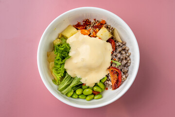 A vibrant bowl brimming with fresh greens, edamame, and cooked grains topped with a rich, creamy dressing. The pastel pink background enhances the colors, inviting a healthy meal experience