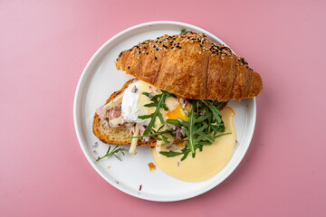 A croissant sandwich showcases layers of savory ham, fresh greens, and a perfectly poached egg, all drizzled with rich hollandaise sauce, creating a delightful breakfast experience
