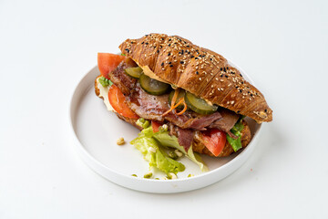 A delightful croissant sandwich bursting with flavors features layers of fresh tomatoes, crisp lettuce, and tender meat. It rests gracefully on a simple white plate that enhances its appeal