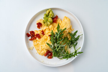A beautifully arranged plate features fluffy scrambled eggs alongside fresh arugula, flavorful sun-dried tomatoes, and creamy avocado chunks