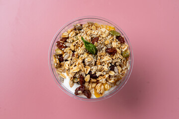 A colorful breakfast bowl features layers of creamy yogurt topped with crunchy granola, dried fruits, and a sprig of mint, creating a dreamy morning treat on a cheerful pink surface