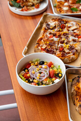 A vibrant salad filled with fresh vegetables sits beside a variety of mouthwatering pizzas atop a rustic wooden table. The ambiance exudes a casual yet inviting spirit, perfect for sharing