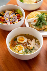 A warm bowl of savory chicken soup with soft boiled eggs sits at the center of a wooden table, surrounded by vibrant salads and comforting pastas, perfect for sharing and enjoying with friends