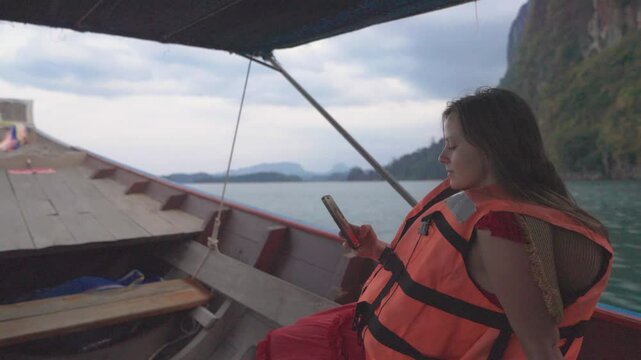 Young female traveler woman is sailing on boat in life jacket. Smiling, he looks camera, then takes a selfie against backdrop of stunning landscapes of Cheo Lan Lake - mountains among turquoise waters