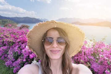 Female traveler in a straw hat takes a selfie against the backdrop of Cheo Lan Lake Dam in Khao Sok...