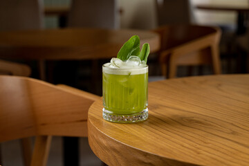 A vibrant green cocktail sits elegantly on a polished wooden table, adorned with fresh herbs and ice, inviting patrons to savor a moment of relaxation in a chic cafe environment