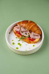 A gourmet croissant sandwich filled with layers of savory ham, crisp lettuce, and vibrant tomato, elegantly presented on a clean white plate. The backdrop features a soothing green tone