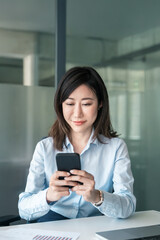 Vertical portrait of mature female businesswoman holding telephone in office for trading, work, looking at screen. 40s mature Asian business woman manager ceo using cellphone mobile phone trade app