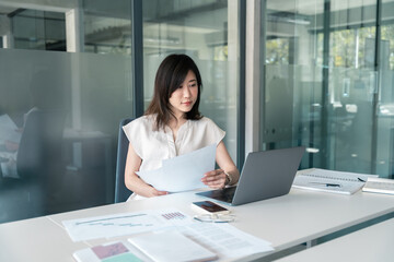 Busy Asian accountant business woman working on laptop computer reading financial document report in office. Middle-aged entrepreneur bank manager businesswoman doing finance paperwork work using pc