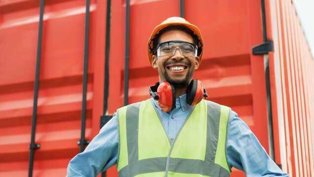 Portrait of cheerful young professional foreman of freight transportation company looking at camera smiling. Happy bearded worker wearing safety helmet, glasses and headphones near red container.