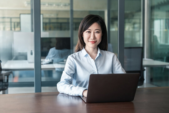 Portrait at camera of 30s it specialist woman in shirt using computer technology for financial bank work online. Mature Asian professional businesswoman working on laptop pc sitting at desk in office - Powered by Adobe