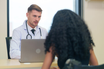 A caucasian doctor in white gown with stethoscope and laptop on table talking to a woman disabled person patient on wheelchair in the examine room