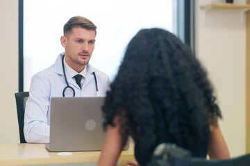 A caucasian doctor in white gown with stethoscope and laptop on table talking to a woman disabled person patient on wheelchair in the examine room