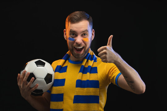 Studio portrait of handsome excited bearded soccer supporter wearing yellow blue t-shirt and scarf is posing with a ball in hand and showing thumbs up gesture, isolated over black background