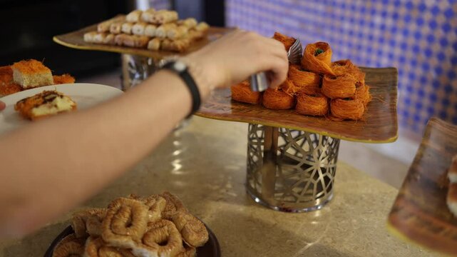 Hand reaching for crispy Middle Eastern sweets kunafa rolls golden layers pistachio filling traditional dessert table luxury buffet textures and flavors rich and inviting festive atmosphere