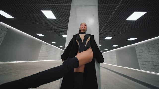 Confident sexy female dancer in black bodysuit, thigh high heeled boots and coat performs stylish choreography in futuristic underground hall tunnel with minimalistic concrete and LED lighting design